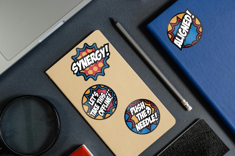 May include: A collection of colorful stickers with bold text and comic-book style designs. The stickers feature phrases like "Synergy!", "Let's Take This Offline!", and "Push the Needle!" on a tan notebook.