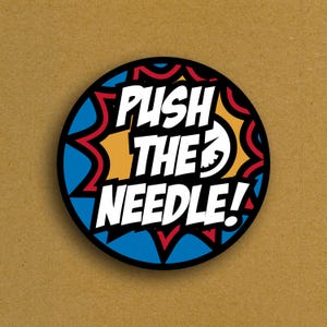 May include: A round sticker with the text "PUSH THE NEEDLE!" in bold white letters. The design features a blue, red, and orange burst pattern around the text, set against a black border. The background is a solid tan color.