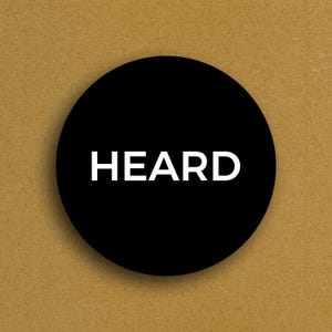 May include: A black circular graphic with the white word "HEARD" centered on a tan background. The image is a simple, bold design, suitable for various applications.