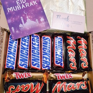 Könnte beinhalten: Eine Geschenkbox gefüllt mit Schokoladenriegeln. Die Box ist mit goldenem Papier ausgekleidet und enthält eine Auswahl an Schokoladenriegeln, darunter Snickers, Twix und Mars. Die Box enthält außerdem eine Karte mit der Aufschrift "Wishing you a very happy Eid Mubarak" und eine Dankeskarte.
