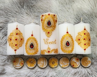 Puede incluir: Conjunto de seis velas blancas con diseños de henna dorados y rojos. Las velas más grandes tienen una sola joya roja en el centro del diseño. Las velas más pequeñas son velas de té. Una de las velas más grandes tiene el nombre "Vincent" escrito en ella.