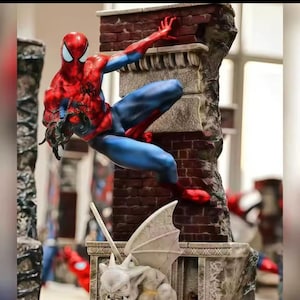 May include: A detailed figurine of Spider-Man in a dynamic pose, clinging to a brick wall. The figure is in a classic red and blue costume, with intricate detailing. The statue is part of a larger display, with other figures visible in the background.