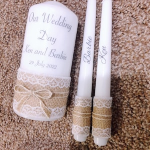 Może przedstawiać: Dwie białe świece ślubne z akcentami z juty i koronki. Większa świeca jest oznaczona napisem "Our Wedding Day Ken and Barbie 29 July 2022". Mniejsze świece są oznaczone napisami "Barbie" i "Ken".