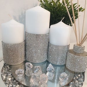 Puede incluir: Tres velas blancas en forma de pilar con bandas de purpurina plateada, una botella de difusor plateada con purpurina y cristales de vidrio transparentes en una bandeja de espejo.