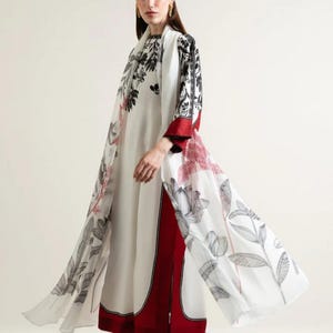 Könnte beinhalten: Ein weißes Kleid und ein passender Mantel mit einem schwarz-roten Blumenmuster. Das Kleid hat einen roten Besatz und der Mantel hat lange Ärmel. Das Outfit wird mit schwarzen Absätzen getragen.