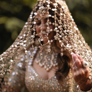 Op de afbeelding: Een gouden bruidssluier met kleine, reflecterende versieringen die over het hoofd en de schouders vallen. De sluier wordt gecombineerd met een bijpassend, rijk versierd lijfje en mouwen, waardoor een samenhangend en elegant ensemble ontstaat. Het ontwerp suggereert een formele of ceremoniële gelegenheid.