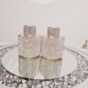 Puede incluir: Dos botellas de vidrio transparente adornadas con brillantes pedrería. Las botellas tienen forma cilíndrica con tapas a juego cubiertas de pedrería. Se exhiben sobre una superficie espejada rodeada de cristales decorativos, reflejando la luz.