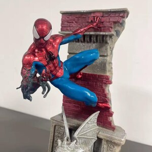 May include: A detailed figurine of Spider-Man in a dynamic pose, clad in a red and blue suit. The figure is set against a backdrop of a brick wall and a gargoyle-adorned structure, capturing a scene of action and adventure.