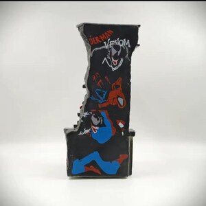 May include: A black, rectangular figurine featuring Spider-Man and Venom graphics. The top of the figurine has the words "Spider-Man Venom" in red and white. The figurine has a jagged edge design and is likely a collectible item.