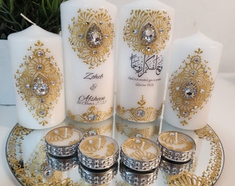 Puede incluir: Velas blancas con detalles dorados y cristales, dispuestas sobre una bandeja de espejo. Las velas presentan diseños ornamentados y texto. También hay velas de té con detalles dorados, creando una exhibición decorativa y elegante.