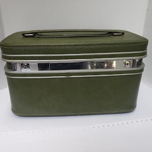 May include: Olive green vintage travel case with a silver metal band and handle. The rectangular case has a rounded top and bottom, with a stitched design around the edges. The case is a classic style, perfect for storing cosmetics or travel essentials.