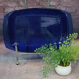 May include: A cobalt blue, rectangular glass dish with a textured pattern, displayed on a silver metal stand. A small white pot with green foliage is to the right of the dish. The dish has a dark blue rim.