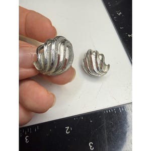 Può includere: Due orecchini in metallo color argento con un design testurizzato a forma di conchiglia vorticosa. L'orecchino più grande è tenuto in mano, mentre quello più piccolo è appoggiato accanto. Un righello nero con segni bianchi indica 2,5 cm.