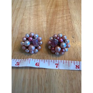 Op de afbeelding: Een paar cluster-oorbellen met diverse tinten roze en witte kunstparels, geaccentueerd met een centrale gefacetteerde roze glaskraal. Een wit meetlint toont een breedte van ongeveer 3,8 cm onder de oorbellen.