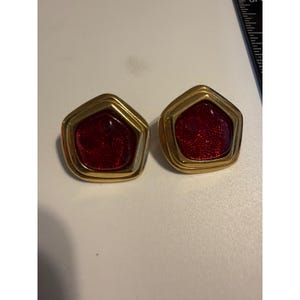 May include: A pair of gold-toned pentagonal earrings featuring a deep red, glittery center inlay. The earrings are positioned next to a black ruler, showing a portion of the measurement scale in inches along the top right edge of the frame.