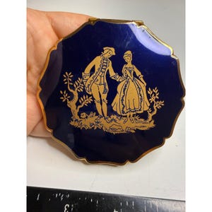 May include: A round, scalloped-edge compact mirror with a deep blue enamel finish and a gold-toned metallic border. The center features a gold-colored illustration of a couple in period attire standing in a landscape with trees.