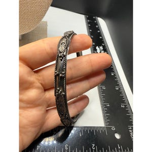 Puede incluir: Un brazalete plateado con intrincados diseños florales y geométricos. El brazalete tiene una superficie texturizada y mide aproximadamente 6,35 cm de diámetro. El diseño incluye elementos florales en relieve y un borde tipo cuerda.