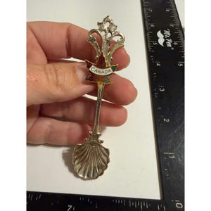 May include: A silver-toned metal souvenir spoon featuring a shell-shaped bowl and a decorative handle with a red and green maple leaf emblem bearing the text CANADA. A black ruler is positioned alongside, showing a length of 4.5 inches.