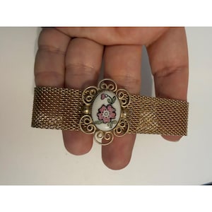 May include: A gold-tone mesh bracelet featuring a central oval-shaped ceramic plaque. The plaque is white with a hand-painted pink flower and green leaves, surrounded by an intricate gold-tone filigree frame with decorative scrollwork.
