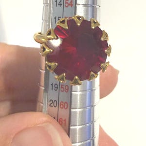 May include: A gold ring featuring a large, round, faceted deep red stone held by a series of small, triangular prongs. The ring is positioned on a silver-colored metal ring sizer tool, with the band resting between the 15 and 16 size markings.