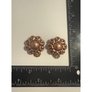 May include: A pair of copper-toned metal clip-on earrings shaped like stylized flowers with a central circular dome and swirling petals. A black L-shaped ruler is positioned next to the earrings, showing each piece is approximately 1.25 inches wide.