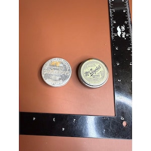 May include: Two round metal snuff tins placed next to an L-shaped black ruler. The left tin is silver with text reading Super-Menthol Singleton's Snuff. The right tin has a cream label with text reading The Original and Genuine McChrystal's Snuff.