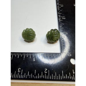 May include: Two small, round, olive green buttons with a carved, ribbed, knot-like pattern. The buttons are positioned next to a black ruler showing measurements in inches, with each button measuring approximately 0.5 inches in diameter.