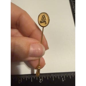 Pode incluir: Um alfinete de lapela dourado com cabeça oval, incrustação de esmalte creme e uma letra A dourada em relevo. O alfinete está ao lado de uma régua preta que mostra um comprimento total de cerca de 6,35 cm de cima para baixo.