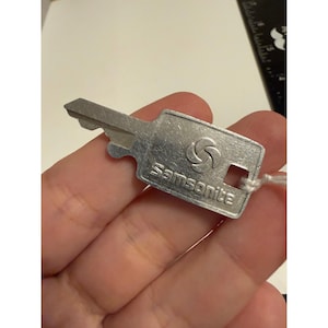 May include: A silver-colored metal luggage key featuring the Samsonite brand logo and name embossed on the head. The key has a standard blade profile with a small hole for a keyring and is shown against a partial view of a black ruler.