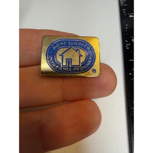 May include: A rectangular gold-tone metal pin featuring a blue oval center with a house icon and the text HOME BUILDERS INSURANCE PROGRAM. The pin is positioned next to a black ruler showing a measurement of approximately 1 inch in width.