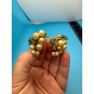 Puede incluir: Un par de pendientes vintage de clip con forma de racimos de uvas. Presentan cuentas de color amarillo crema con aspecto de perlas, hojas de esmalte verde y detalles metálicos dorados. Los pendientes se sostienen en una mano sobre un fondo azul.