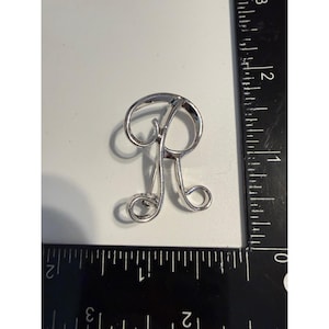 Puede incluir: Un broche de metal plateado con la forma de la letra R cursiva. El broche está colocado junto a una regla blanca y negra, mostrando que el artículo mide aproximadamente 3,8 cm de alto y 3,2 cm de ancho.