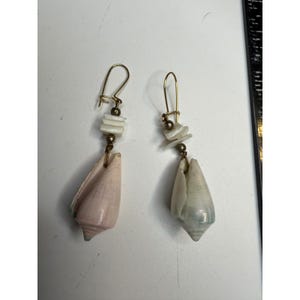 Puede incluir: Un par de pendientes colgantes con ganchos de alambre dorado, pequeñas cuentas metálicas y espaciadores rectangulares de concha blanca. Cada pendiente termina en una concha cónica, una de color rosa suave y la otra de color azul grisáceo.