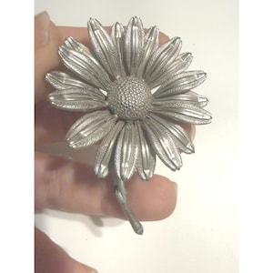 Puede incluir: Un broche de metal plateado con forma de flor de margarita. La pieza presenta un centro texturizado y punteado rodeado de pétalos alargados y ranurados que irradian hacia afuera. Un tallo de metal delgado y curvo se extiende desde la base.