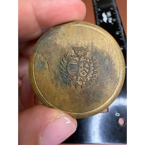 May include: A round, brass-toned metal compact featuring a fine, vertical-lined texture and a central engraved crest with a crown and shield motif. A black ruler is visible in the background, showing the object is approximately 2 inches wide.