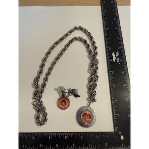 Può includere: Una collana a catena ritorta color argento con un ciondolo circolare raffigurante una fotografia di due persone, accompagnata da una spilla abbinata con design a fiocco e un ciondolo fotografico circolare più piccolo. Un righello indica 30,5 cm.