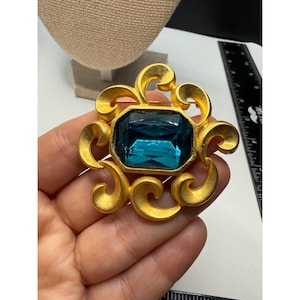 Puede incluir: Broche dorado con una gran gema rectangular de color turquesa en el centro. La gema está rodeada por un diseño dorado ornamental y arremolinado. Un accesorio vintage.