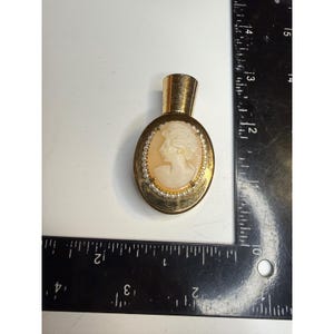 May include: An oval gold-tone perfume bottle featuring a central cameo of a profile portrait surrounded by a border of small white beads. The bottle is positioned next to a black L-shaped ruler, showing a height of approximately 2.5 inches.