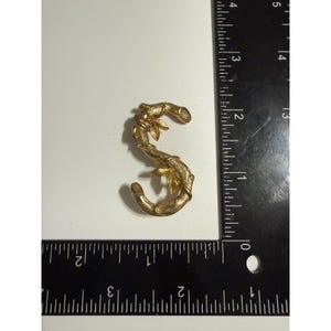 May include: A gold-toned metal brooch shaped like the letter S, featuring a textured bamboo-like design with small leaves. The brooch is positioned next to a black L-shaped ruler, showing the piece measures approximately 6.4 cm in height.