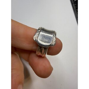 May include: A silver-toned spoon ring featuring a wide, smooth, rectangular center panel with rounded corners and decorative embossed scrollwork along the edges. The band is formed from the handle of a spoon, showing a tapered, textured design.