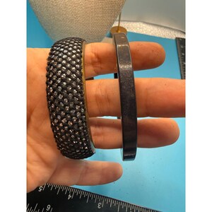 May include: Two bracelets. One is black with a textured surface of small, shiny embellishments. The other is a solid, dark gray, smooth band. The bracelets are held in a hand against a blue background.