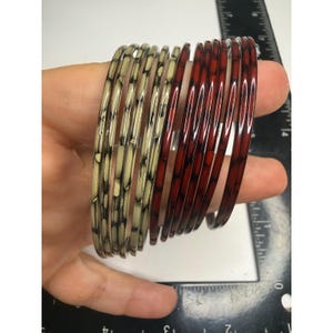 May include: A set of thin bangles featuring a mottled pattern in cream and dark red colors. The bangles are positioned next to a black ruler showing measurements in inches, with the bangles measuring approximately 2.5 inches in diameter.