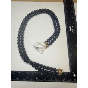 May include: A double-strand necklace featuring smooth black beads and a central white rose-shaped pendant with gold-tone accents. A black ruler is positioned along the side and bottom, showing the necklace length is approximately 40.6 cm.