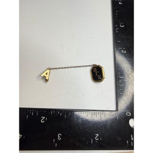 May include: A gold-toned pin featuring the letter A connected by a thin metal chain to an octagonal black and gold badge displaying the Greek letters Beta, Gamma, and Chi. A black ruler with white markings is visible, showing a length of 5 cm.