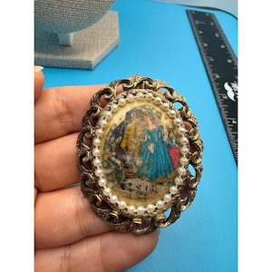 May include: Vintage oval brooch with a detailed image of figures in a garden scene. The brooch has a gold-tone filigree frame, a border of small faux pearls, and a light-colored background. The brooch is approximately 2 inches in length.