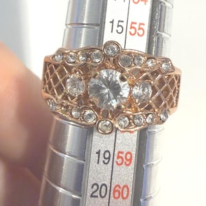 May include: A rose gold-toned ring featuring a large central clear stone flanked by two smaller clear stones, surrounded by a lattice pattern and additional small clear stones, positioned on a metal ring sizer showing a size between 19 and 20.