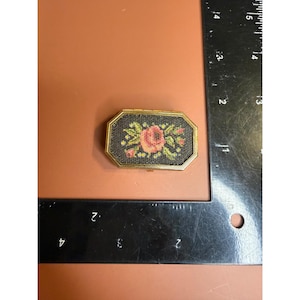 May include: A small rectangular gold-toned metal box with a needlepoint floral inlay featuring a pink rose and green leaves on a black background, positioned next to a black metal ruler showing a width of approximately 1.5 inches.