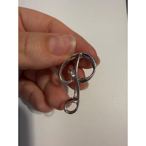 May include: A silver-toned metal brooch shaped like the cursive letter P. The design features a continuous, polished loop with a swirling flourish at the top and a rounded base. The surface is smooth and reflective, showcasing a metallic finish.