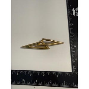 May include: A gold-toned metal brooch featuring an abstract, elongated geometric design composed of two interlocking triangular shapes. The brooch is positioned next to a black ruler, showing a total length of approximately 3.5 inches.