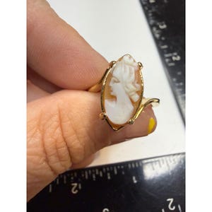 May include: A gold-toned ring featuring an oval-shaped cameo with a white profile carving set against a light brown background. The cameo is held by four prongs on a simple band, positioned next to a black ruler showing metric measurements.
